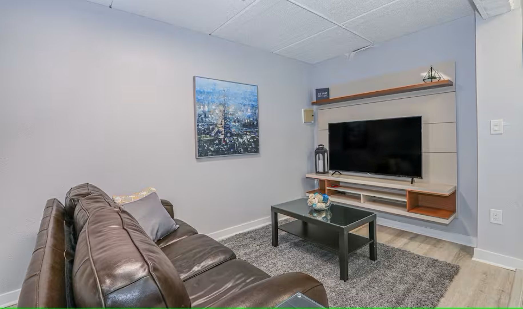 A living room with a leather sofa, a coffee table, a flat-screen TV, a framed picture on the wall, and a small shelf above the TV.