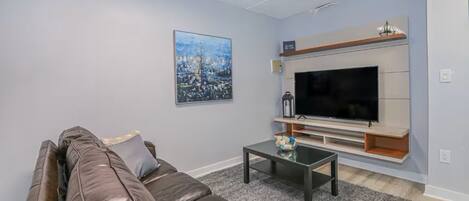 A living room with a leather sofa, a coffee table, a flat-screen TV, a framed picture on the wall, and a small shelf above the TV.