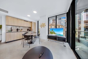 Une cuisine moderne avec des meubles en bois, un coin repas avec une table ronde et des chaises, un grand téléviseur à écran plat et un balcon offrant une vue sur la ville.