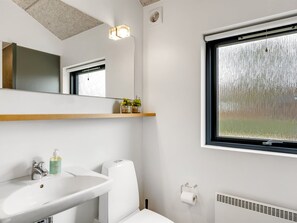 A modern bathroom with a white sink, toilet, and window. There is a shelf with plants and a mirror above the sink. - "Anttoni" - 350m from the sea by Interhome (Sæby)