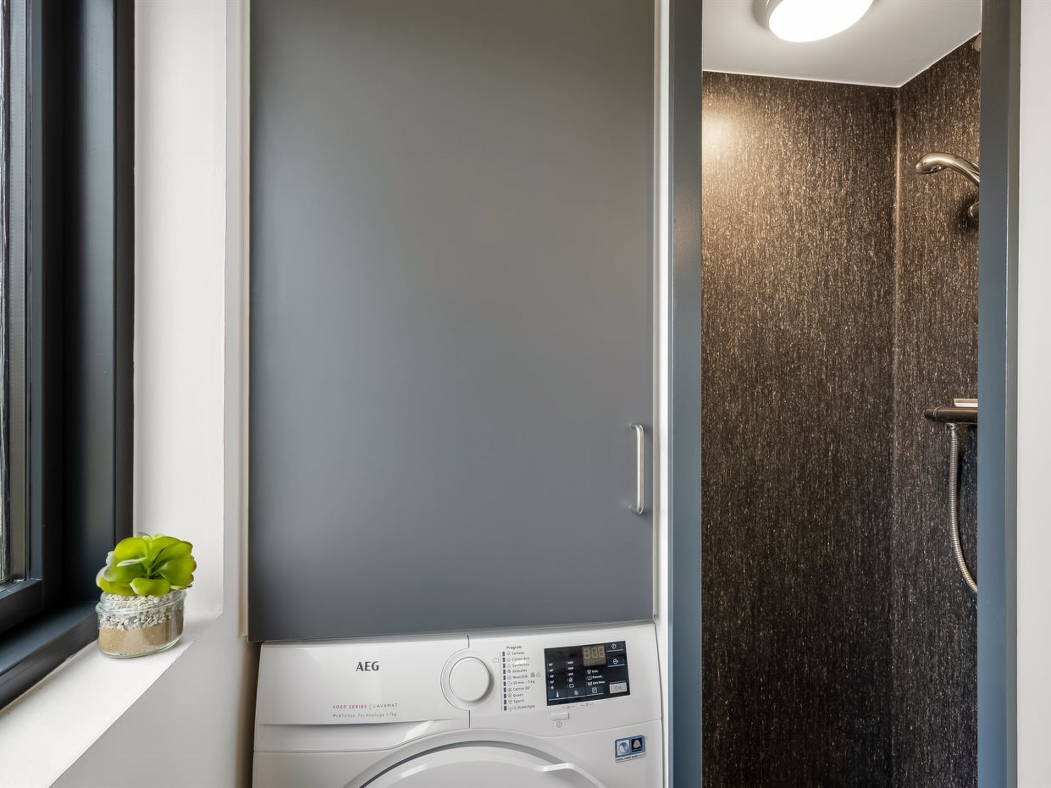 Ein kleines, modernes Badezimmer mit integrierter Waschmaschine und Trockner, einer kleinen Topfpflanze und einer Duschkabine.