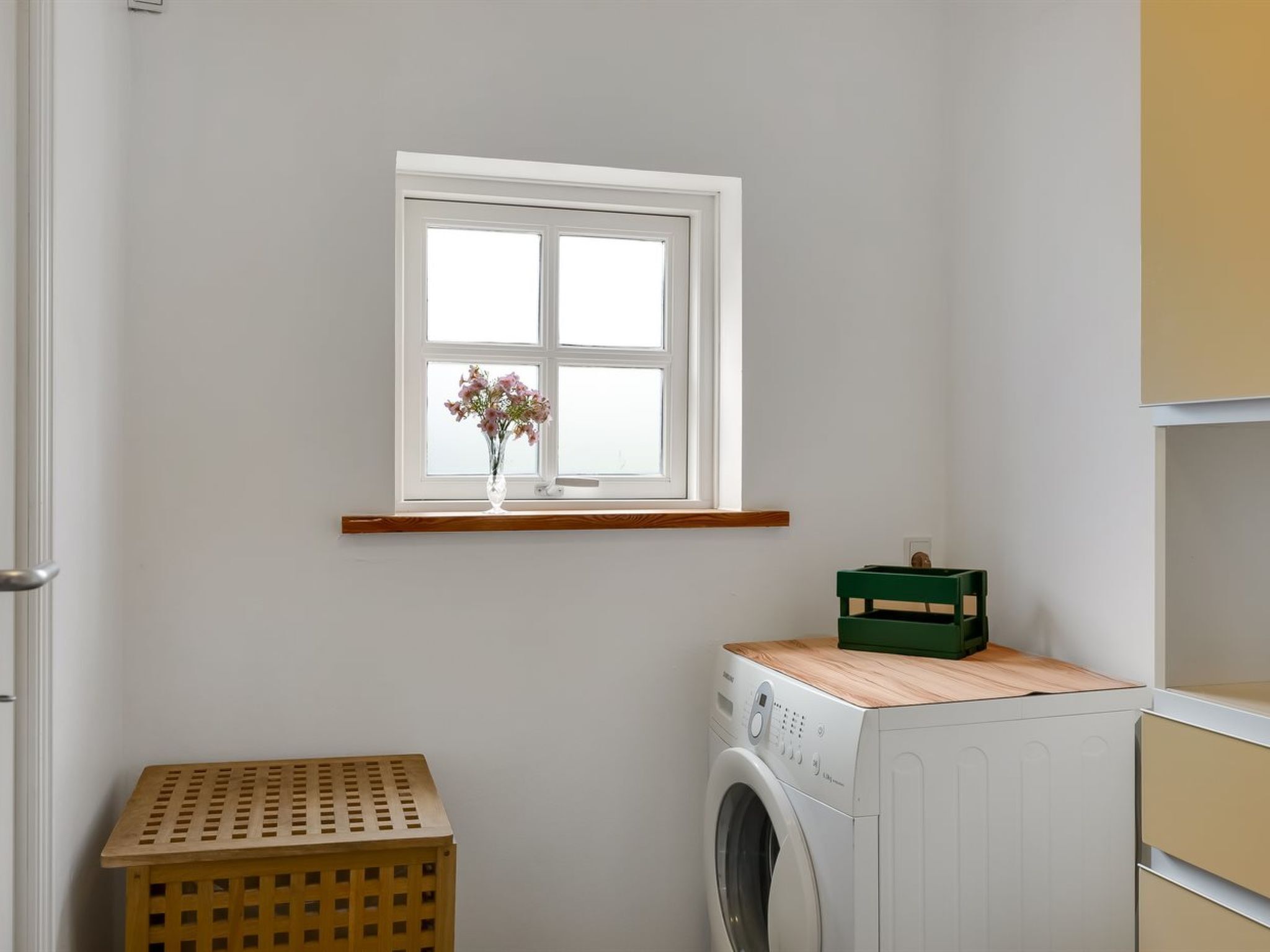 Een wasserette met een wasmachine, een houten kist, een groene doos en een raam met een bloevas.