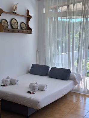 Ein ordentlich bezogenes Bett mit Kissen, eine kleine Ablage mit dekorativen Tellern und Blick durch leichte Vorhänge.