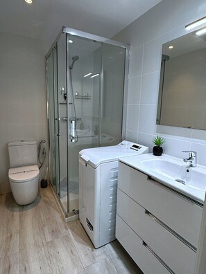 Un baño moderno con ducha de vidrio, inodoro, lavadora y lavamanos con espejo.