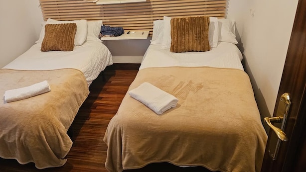 Une chambre d’hôtel avec deux lits, chacun recouvert d’une couverture marron et doté d’un oreiller blanc. Le sol est en bois et la porte a une poignée dorée.