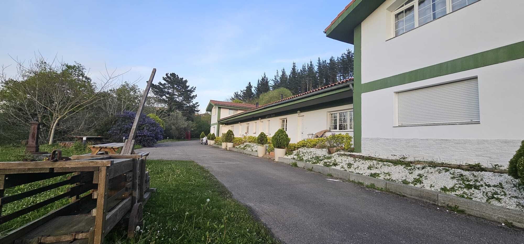 A building with a green and white facade, a paved driveway, and a grassy area with trees and shrubs.