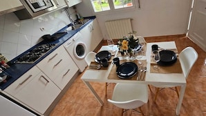 Une cuisine compacte équipée d’une cuisinière à gaz, d’un lave-linge et d’une table à manger pour deux personnes, avec assiettes, couverts et verres.