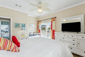 Ein Schlafzimmer mit Bett, Deckenventilator, Fernseher, Kommode und einem Balkon mit Vorhängen.