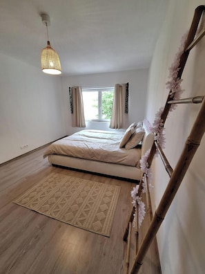 A bedroom with a bed, a hanging light fixture, a window with curtains, and a wooden floor.