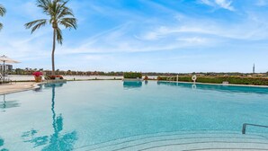A clear swimming pool with a palm tree, a lifeguard stand, and a distant view of hotel buildings.