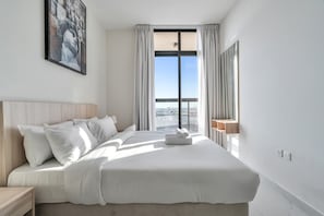 A modern hotel room with a large bed, a nightstand, a painting, a window with curtains, and a view of the city. - Free Housekeeping Visits During Your Stay - Stayshort - Comfortable 1 Bed in Jebel Ali Sleeps 2 With Balcony (Dubai)