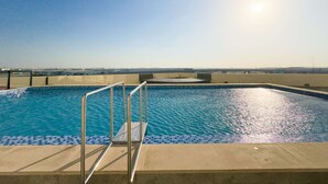 Una piscina con escalera de metal, agua azul cristalina y vista a un horizonte urbano lejano bajo un cielo soleado.