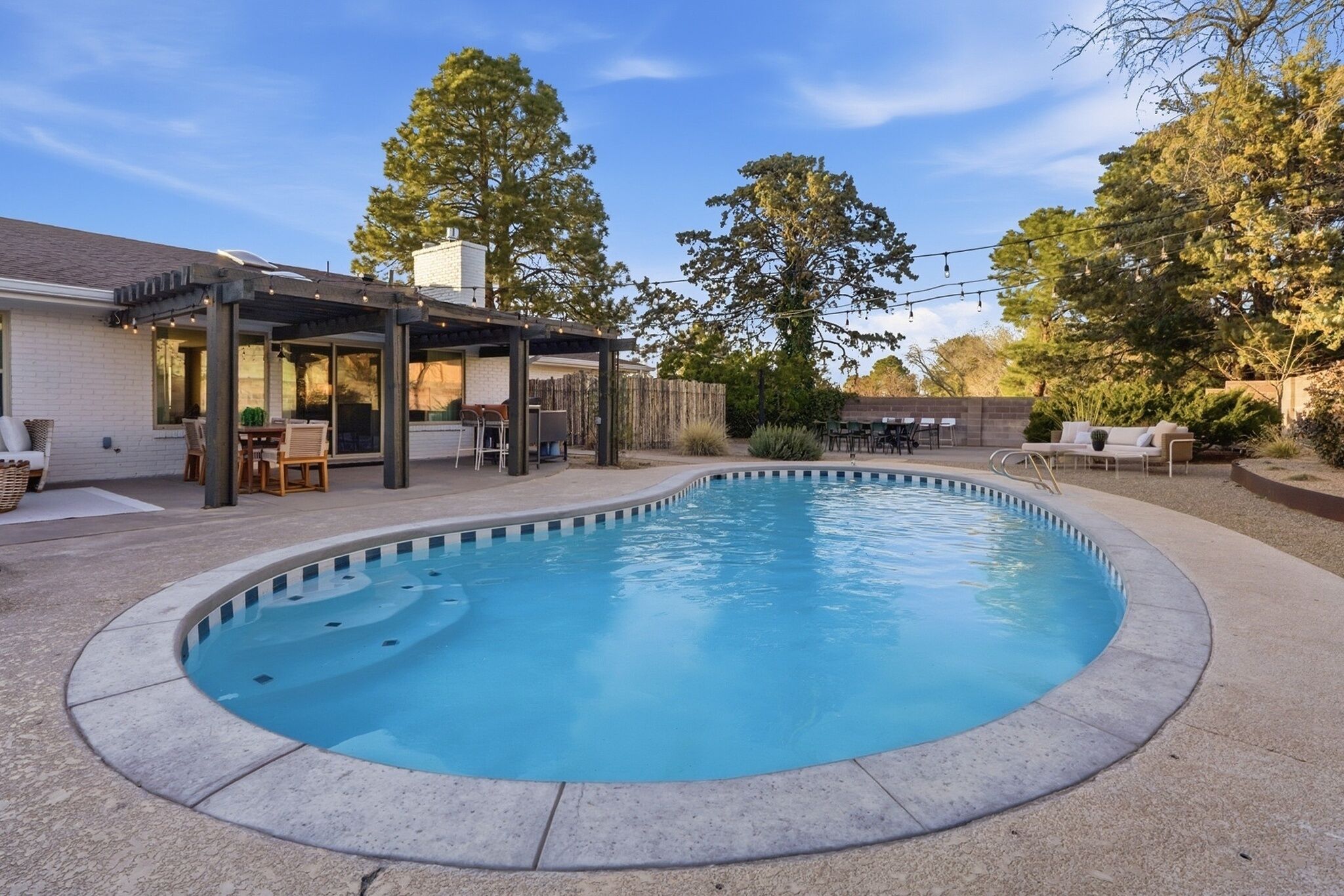 A swimming pool with a covered patio area, outdoor furniture, and trees in the background.