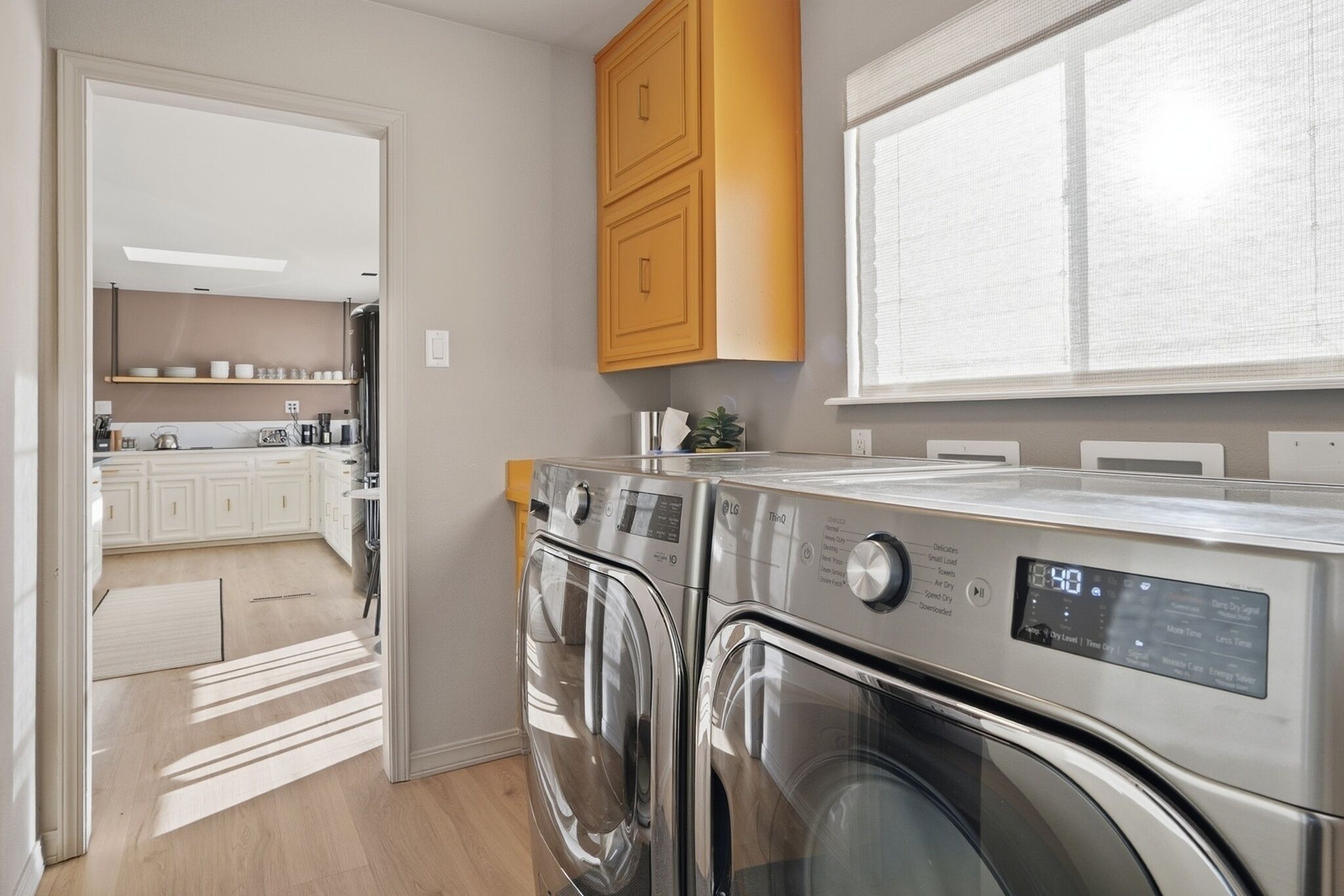 A laundry room with two front-loading washing machines, a wooden cabinet above, and a view of a kitchen area through an open door.