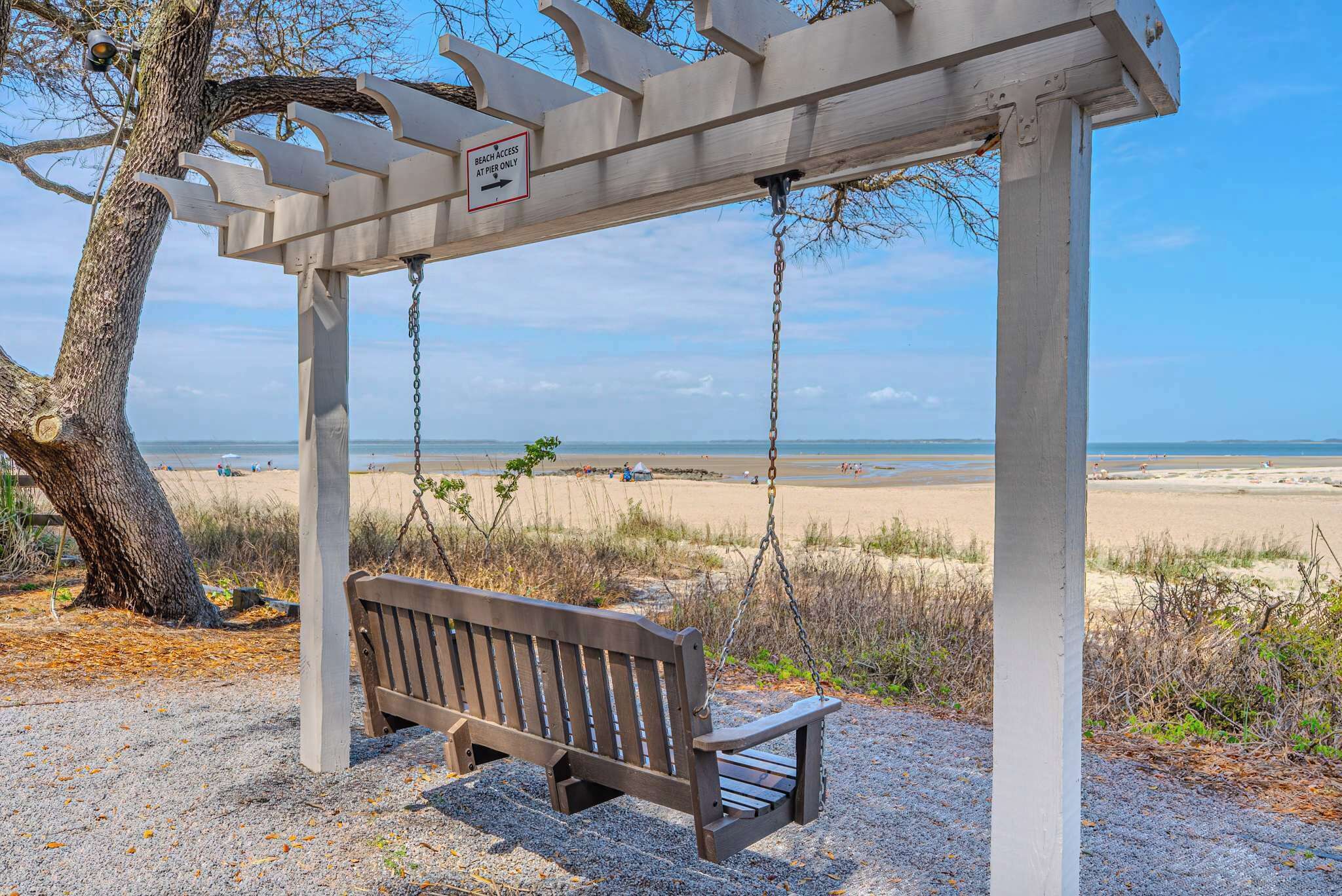 A wooden swing bench under a white pergola with a beach view.