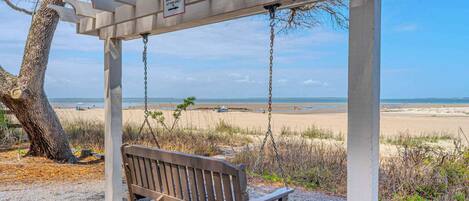 A wooden swing bench under a white pergola with a beach view.