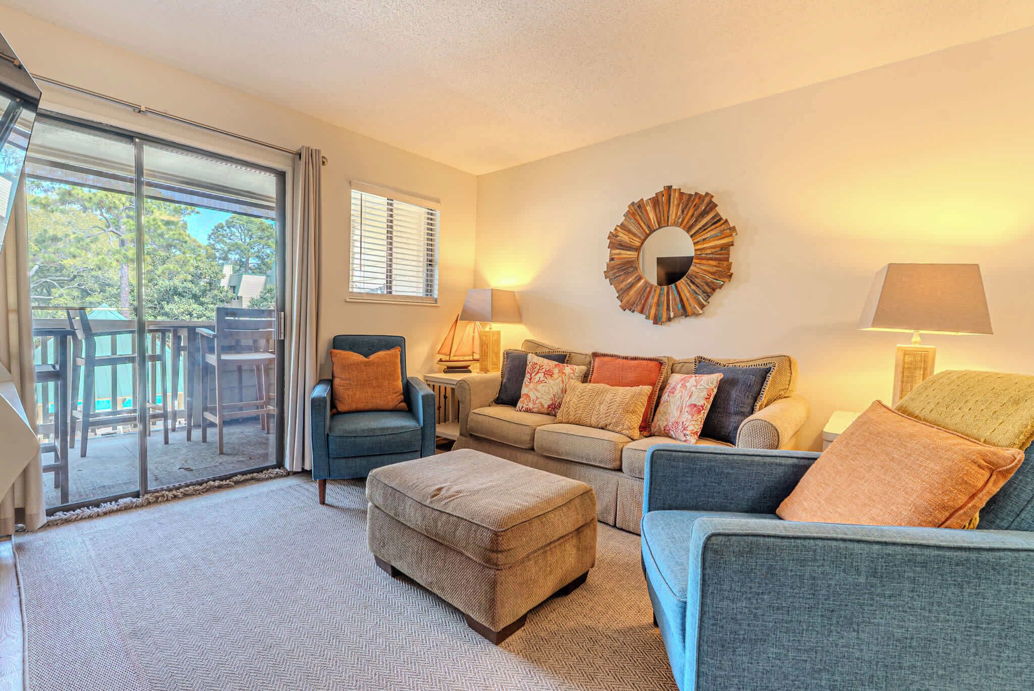 A living room with a sofa, ottoman, and two chairs, a coffee table, a lamp, a mirror, and a sliding glass door leading to an outdoor area.