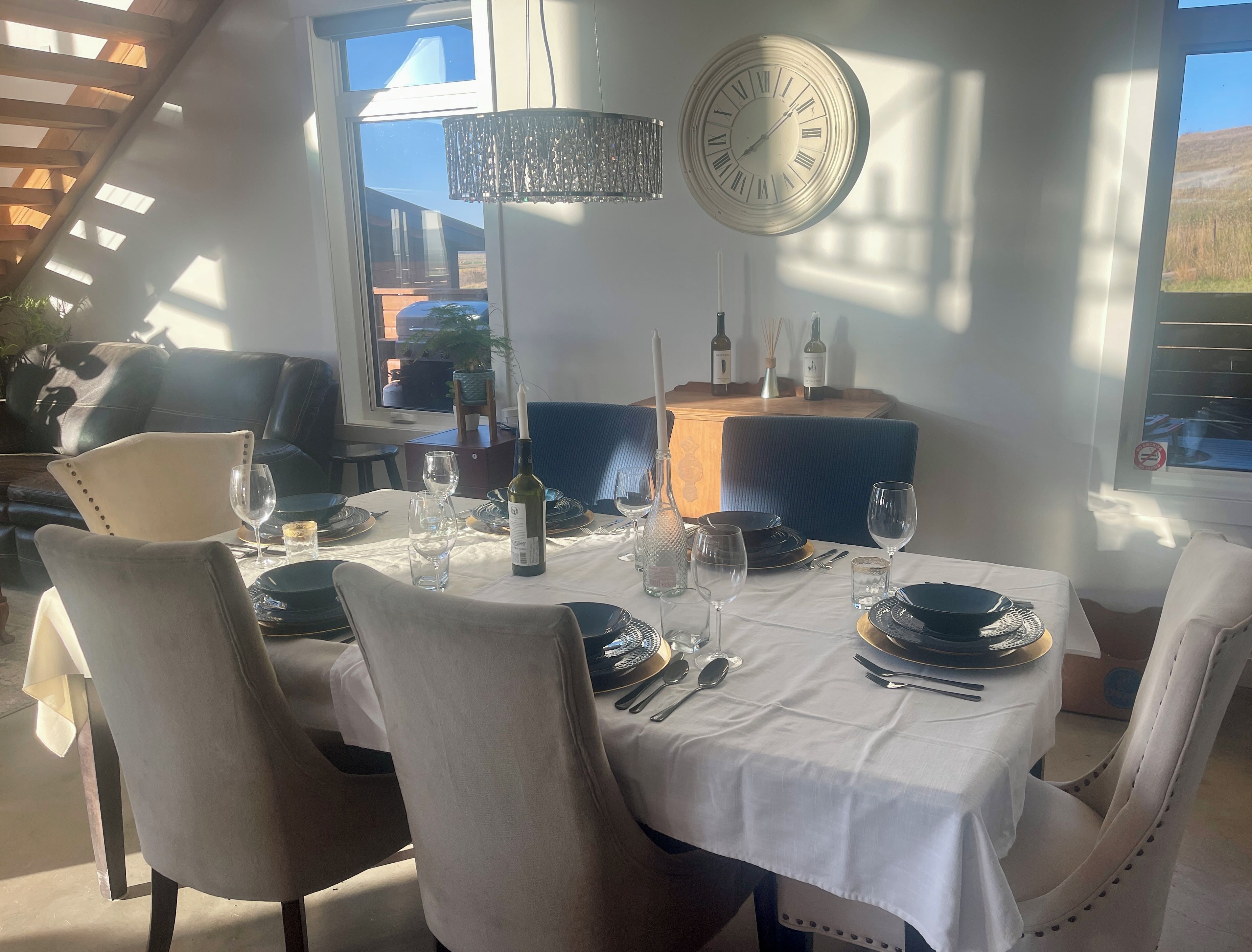 A dining area with a table set for two, a clock on the wall, and a view of the outdoors.