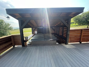 A covered outdoor seating area with a hot tub, wooden deck, and a view of the countryside.