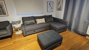 Ein modernes Wohnzimmer mit einem grauen Ecksofa, passendem Hocker, einem kleinen weißen Beistelltisch und gerahmten Kunstwerken an den Wänden.
