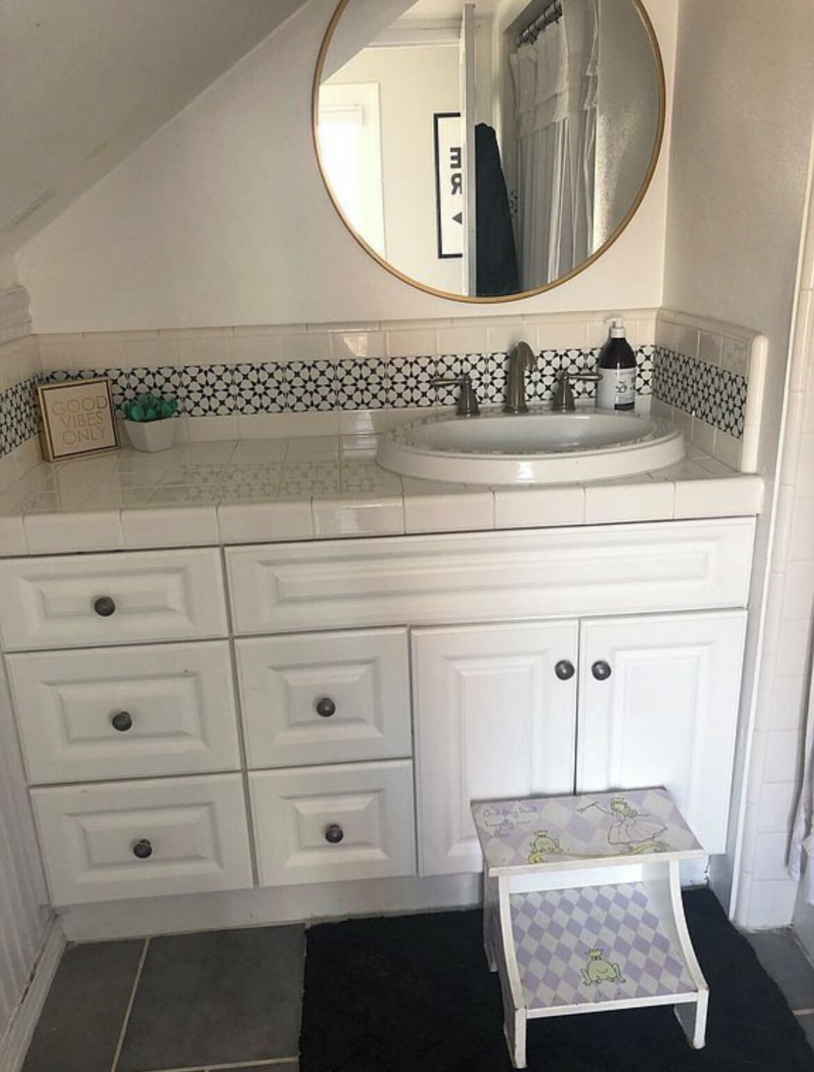 A bathroom with a white vanity, a round mirror, a sink, and a small stool.