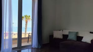 Ein Zimmer mit einer Schiebeglastür, die Blick auf einen Strand bietet, einem dunkelgrauen Sofa mit Kissen und einem Sessel an der Tür.