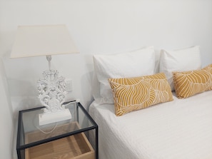 A bed with white and patterned pillows, a white bedside table with a lamp, and a glass side table with a wooden tray. - El Rincon de San Fermin (Zizur Mayor)