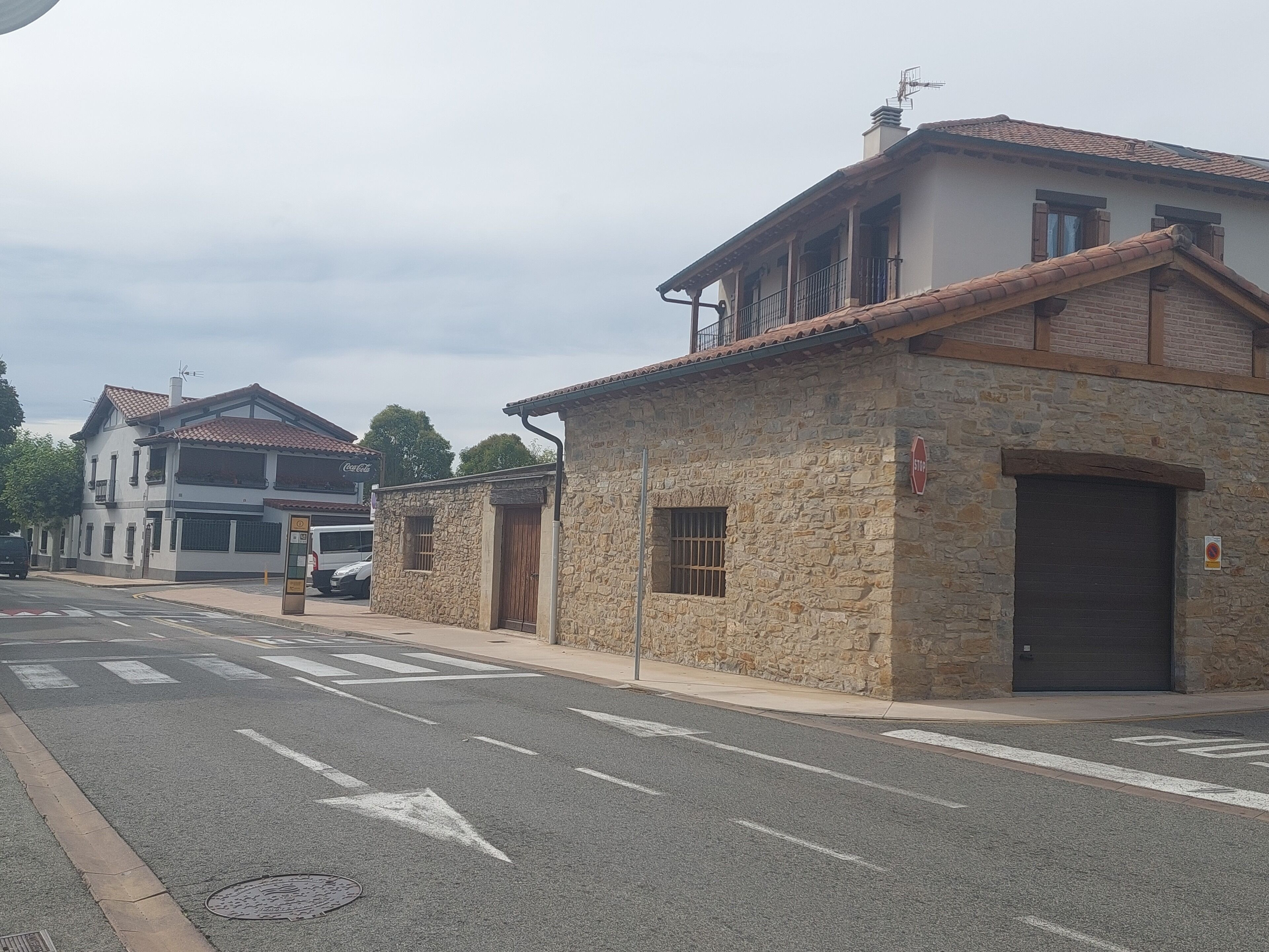 A street with buildings, including a stone building with a closed shutter and a two-story building with a tiled roof.