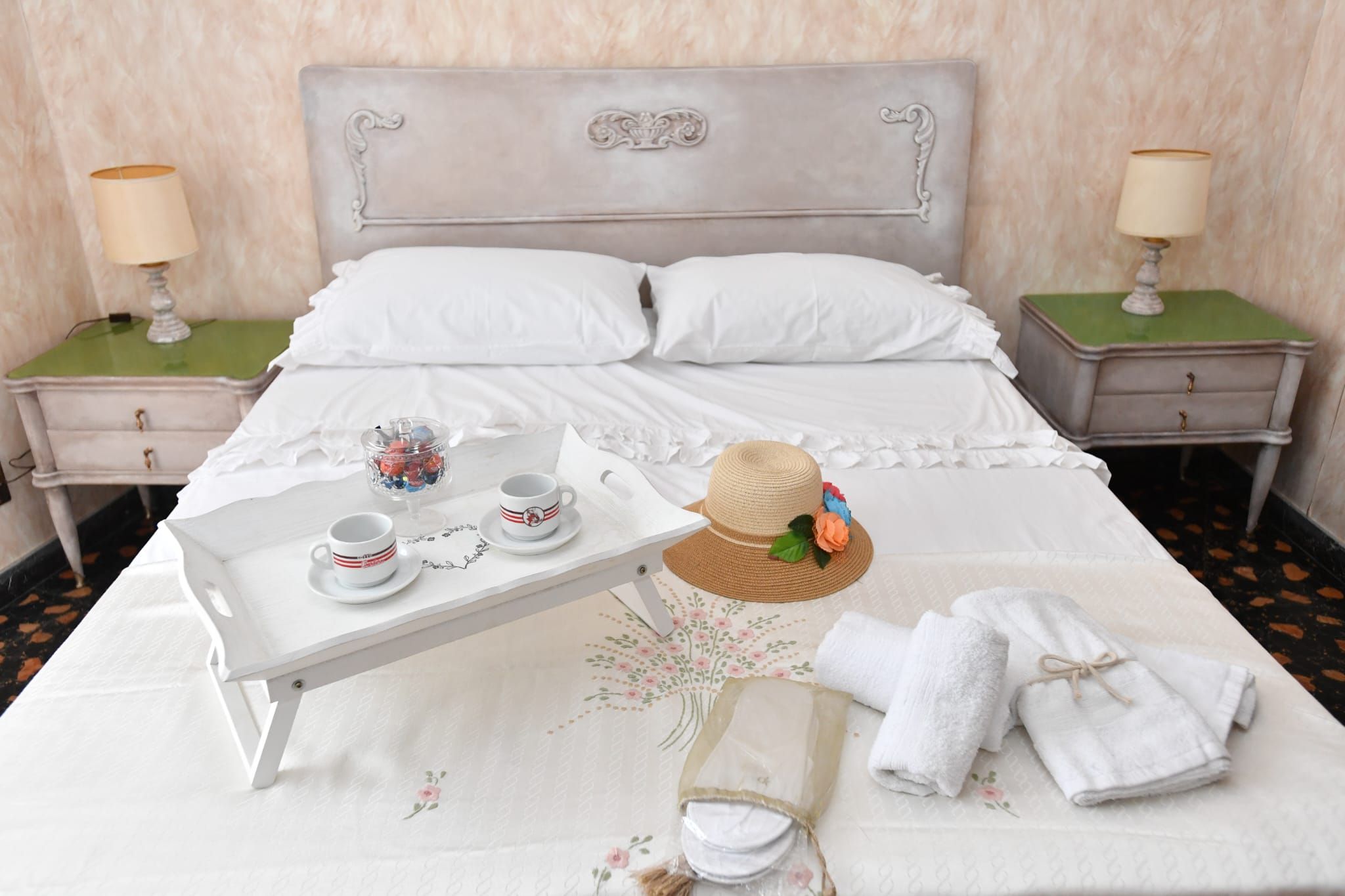 Una cama bien hecha con sábanas blancas, una bandeja con tazas y un sombrero, y toallas sobre la cama.