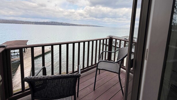 Een balkon met uitzicht op een meer, een houten terras en twee rieten stoelen.