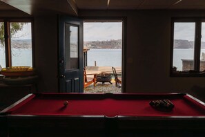 A pool table with a red felt surface, a door leading outside, and a view of a lake and distant buildings. - Lakeside Retreat with Stunning Waterfront Views (Rushville)