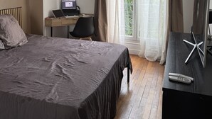 Ein Schlafzimmer mit einem Bett, einem Schreibtisch mit Laptop, einem Fernseher und einem Deckenventilator.