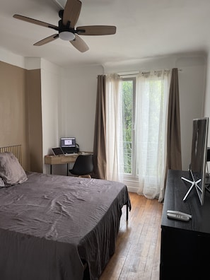 Une chambre à coucher avec un lit, un bureau avec un ordinateur portable, une télévision et un ventilateur de plafond.