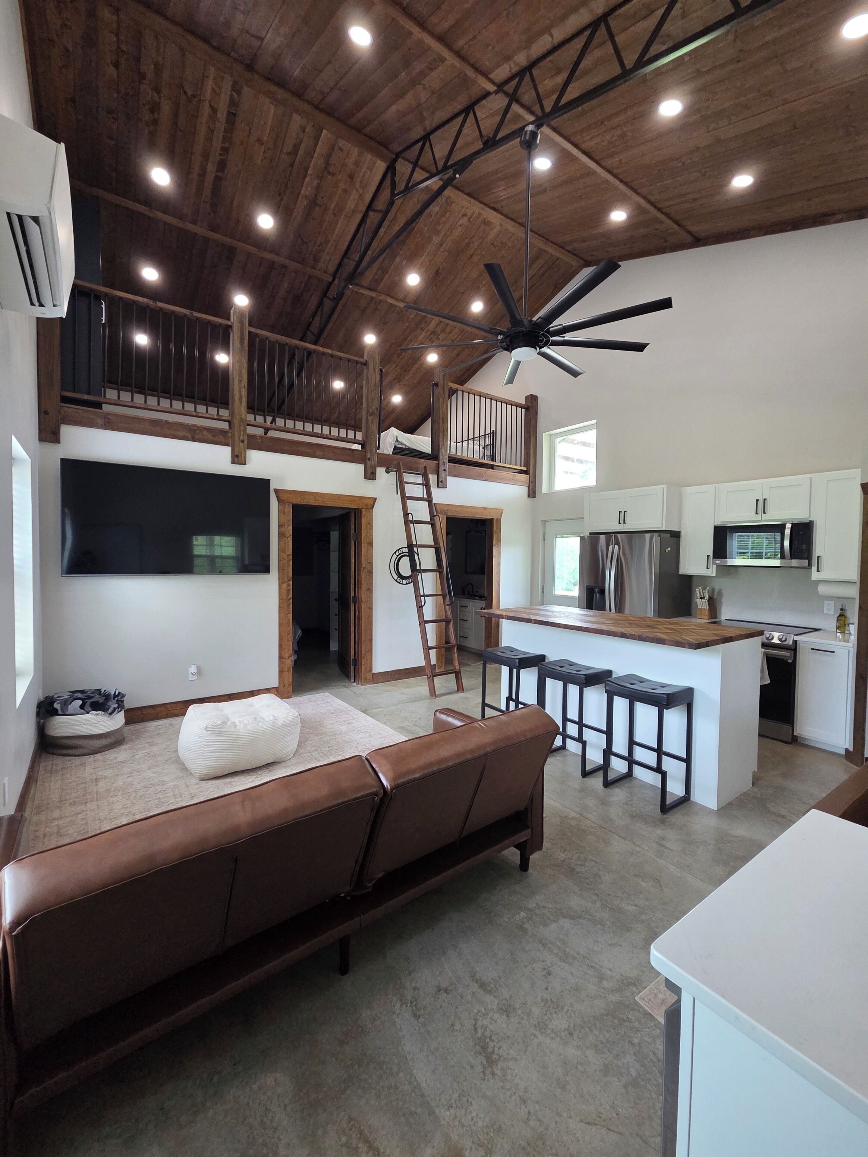 Un vaste espace de vie doté d’un haut plafond, une chambre en mezzanine, une cuisine équipée d’appareils électroménagers modernes et un coin salon avec un canapé en cuir marron.