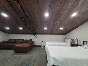 A room with a sloped ceiling, wooden beams, a brown sofa, two beds with white bedding, and a small table with a lamp.