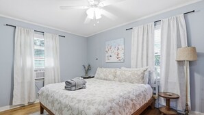 Un dormitorio con cama, ventilador de techo, mesita de noche, lámpara y ventana con cortinas.