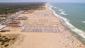 Ein Strand mit Reihen von Sonnenschirmen und Liegestühlen, einem Parkplatz und Gebäuden im Hintergrund.