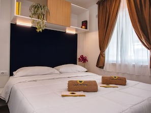A hotel room with a bed, a nightstand, a shelf with books and a plant, and a window with curtains. - Tenuta Primero Grado Family Collection (Grado)