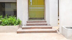 A building entrance with a glass door, steps leading up to it, and a small planter with green plants.