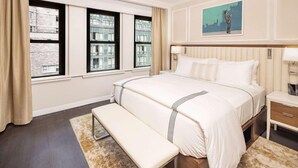 Quarto de hotel com uma cama grande, um banquinho, uma mesinha de cabeceira e um quadro na parede.