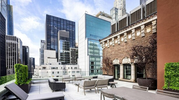 Um terraço no telhado com área de estar externa, um edifício de tijolo com arquitetura clássica e edif ícios altos modernos ao fundo.