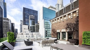 A rooftop terrace with outdoor seating, a brick building with classical architecture, and modern high-rise buildings in the background.