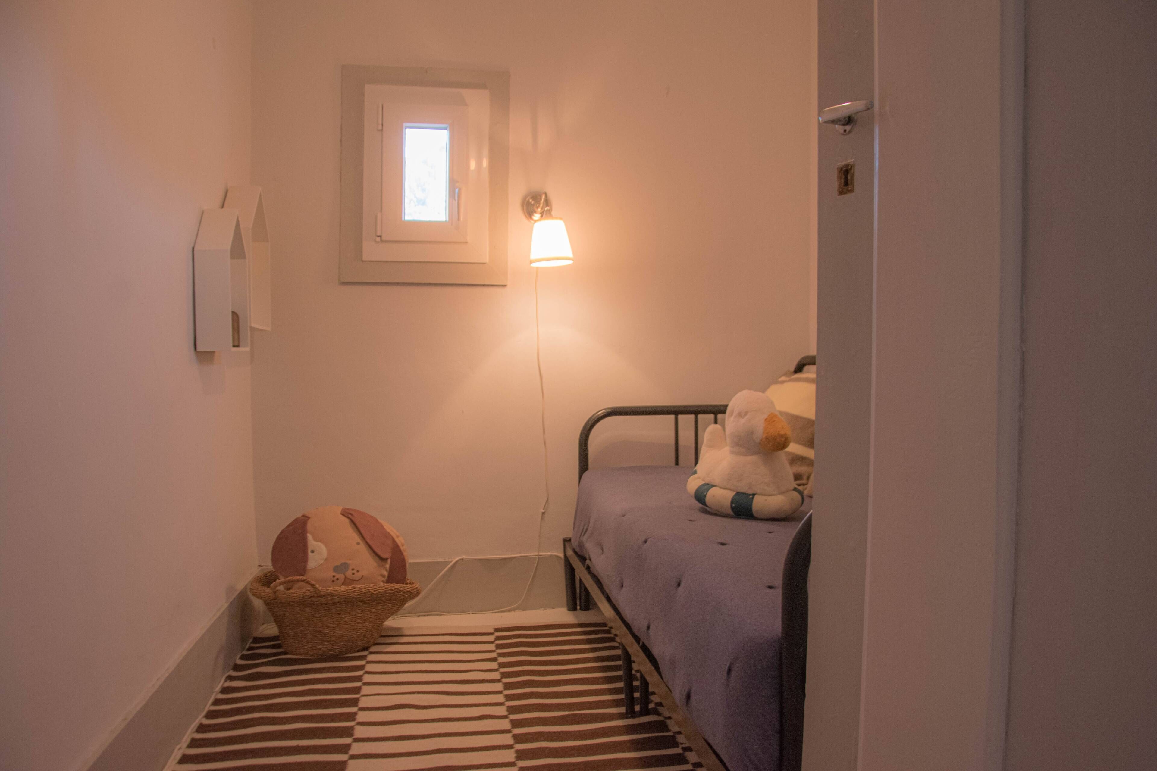 Ein kleiner, schmaler Raum mit einem Einzelbett, einem Nachttisch, einer Lampe, einem Fenster und einem Wandregal.