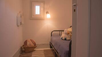 Ein kleiner, schmaler Raum mit einem Einzelbett, einem Nachttisch, einer Lampe, einem Fenster und einem Wandregal.