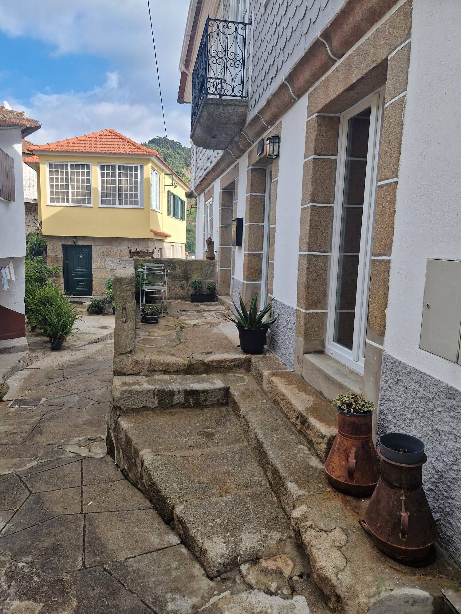 Un étroit sentier de pierre mène à un bâtiment à la façade blanche et au toit recouvert de tuiles rouges. On y trouve des plantes en pot et une chaise en métal à l’extérieur.