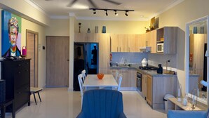 A modern kitchen with wooden cabinets, a white countertop, and a dining area with a blue chair and a white table. - Ocean View | Central SD | Rooftop Plunge Pool (Santo Domingo)