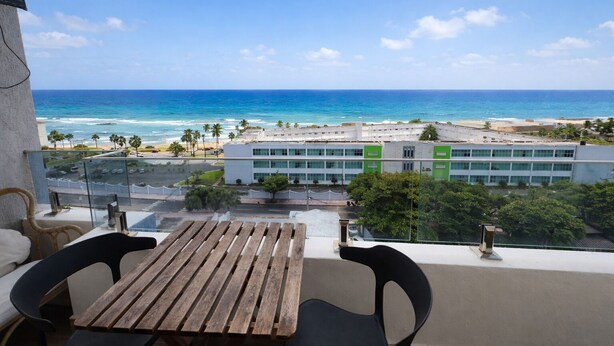 A balcony with a wooden table and chairs, overlooking a beach and a building with large windows. - Ocean View | Central SD | Rooftop Plunge Pool (Santo Domingo)