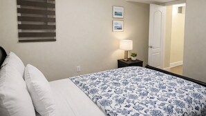 A hotel room with a bed, a nightstand with a lamp, and a door leading to another room. - Cozy 2BR Basement | Private Entry + Parking (Brampton)