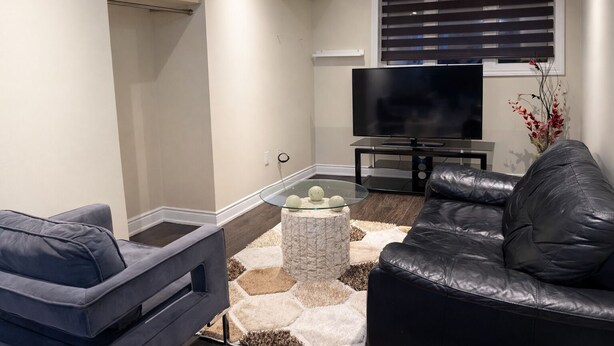 A modern living room with a sofa, a glass coffee table, a television, and a window with blinds. - Cozy 2BR Basement | Private Entry + Parking (Brampton)