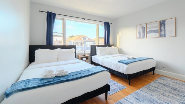 Habitación de hotel con dos camas, una ventana con cortinas, una mesita y una obra de arte colgada en la pared.